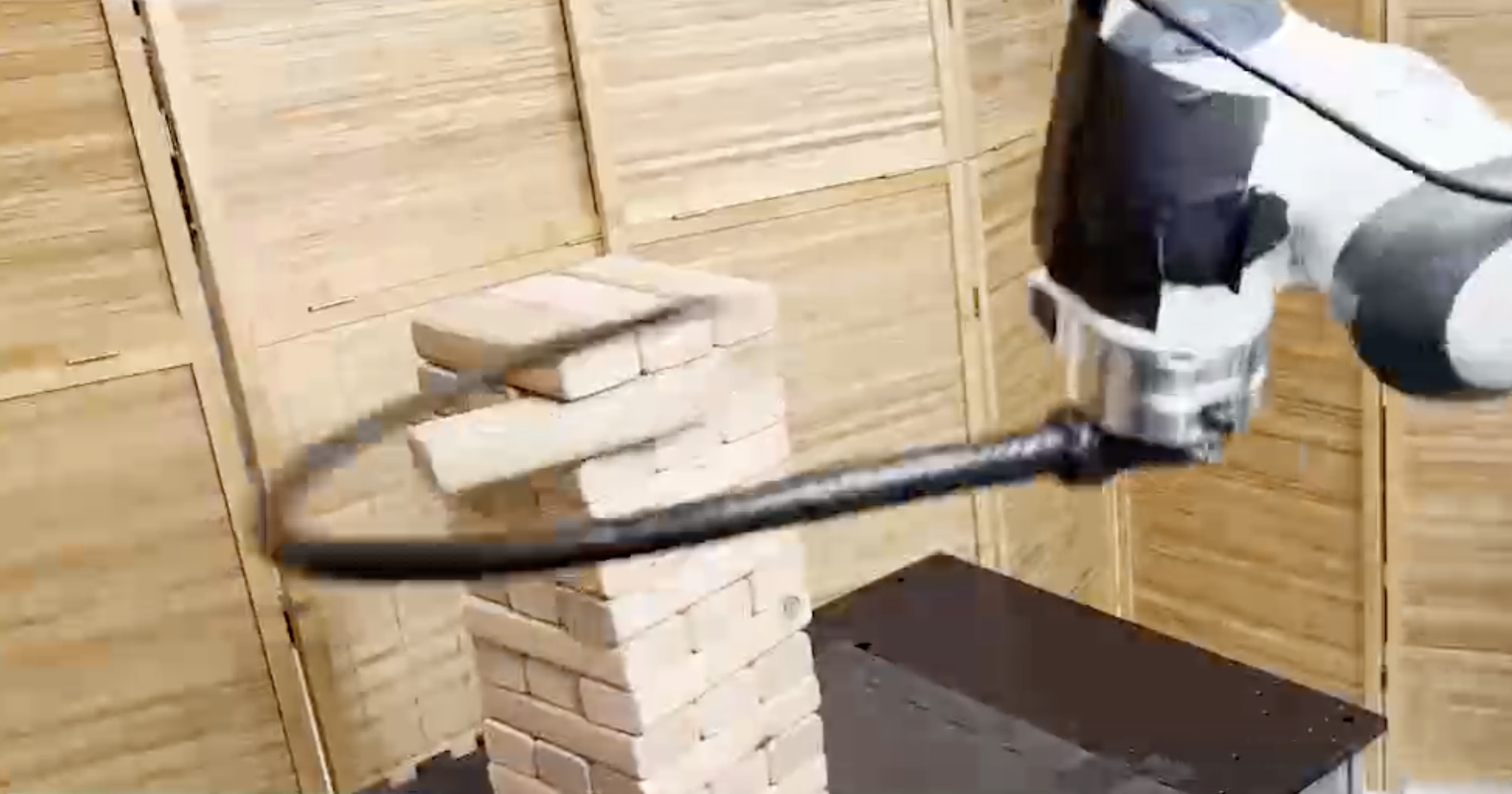 a black-and-silver robotic arm with a black whip attached has just struck a tower of Jenga blocks. One of the blocks is partially dislodged and about to fall out from the tower.